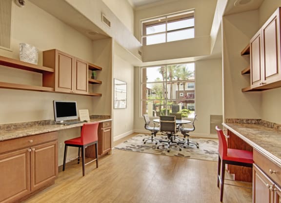 Modern office space here at Mozaic at Steele Park Apartments with wooden floors, large window, two workstations with computers, red chairs, and a round meeting table with rolling chairs. Bright and airy ambiance.