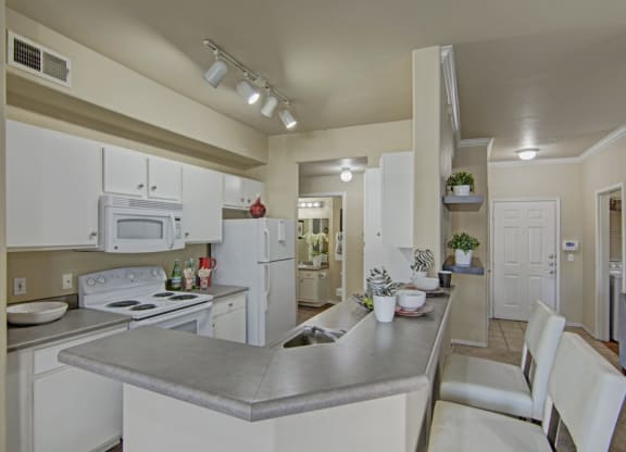 Modern kitchen here at Mozaic at Steele Park Apartments with white cabinets, gray countertops, and bar seating. Bright lighting, decorative plants, and a cozy, welcoming ambiance.