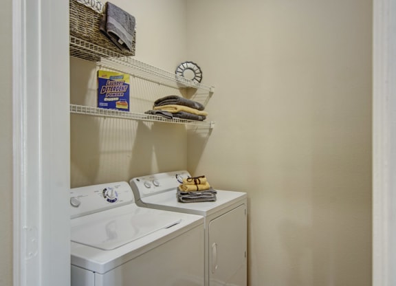 Compact laundry room here at Mozaic at Steele Park Apartments with a washing machine and dryer. Shelves hold detergent, a wicker basket, and neatly folded towels, creating an organized feel.