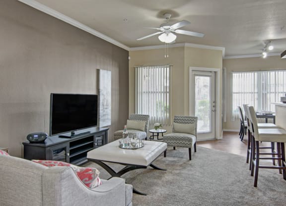 Modern living room here at Mozaic at Steele Park Apartments with neutral tones, featuring a TV on a stand, a gray sofa, patterned chairs, bar stools, and tall windows letting in natural light.