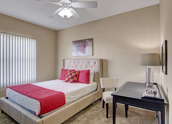 A cozy bedroom here at Mozaic at Steele Park Apartments with a beige tufted bed, white and red bedding, and a checkered chair. A black desk with a lamp and modern decor adds elegance.