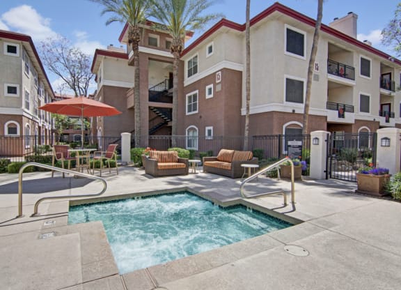 Sunny courtyard with bubbling hot tub, surrounded by metal railing. Two-story beige Mozaic at Steele Park Apartment building, palm trees, and patio chairs create a relaxing vibe.