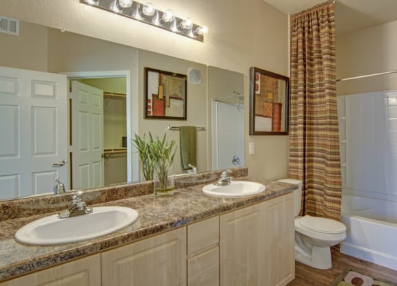 Modern bathroom here at Natura VIllas Apartments with dual sinks on a marble countertop, surrounded by beige walls. A leafy plant, abstract art, and a patterned shower curtain add decor.
