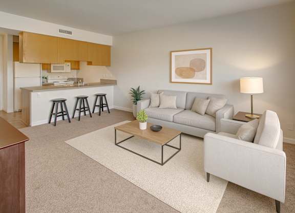 A bright, modern living room here at Confluence at Harvest Hills featuring an open-concept layout with a neutral sofa and armchair, a wooden coffee table on a light area rug, soft beige carpeting, a kitchen breakfast bar with stools, light wood cabinetry, a floor lamp, simple abstract wall art, and large windows allowing natural light to fill the space.