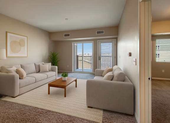 A bright living room here at Dakota Commons featuring two light neutral sofas arranged across a wooden coffee table, a soft area rug over carpeted flooring, neutral wall art, and a floor lamp, with sliding glass doors leading to a private balcony that brings in natural light and offers an open, comfortable layout.