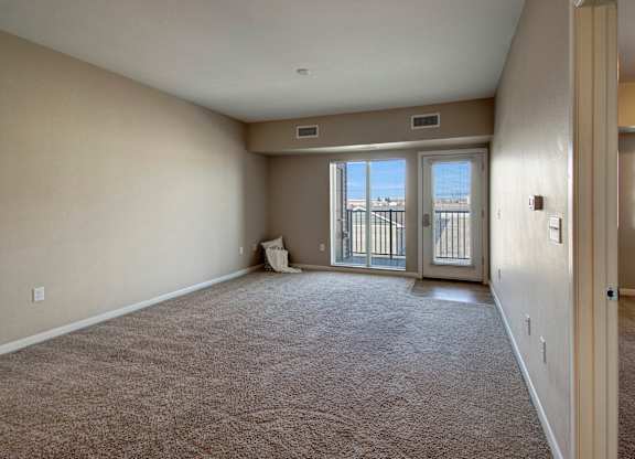A spacious, unfurnished living room here at Dakota Commons featuring soft neutral walls, plush carpet flooring, and a sliding glass door with an adjacent exterior door that opens to a private balcony, allowing natural light to fill the open layout and highlight the room’s generous size.