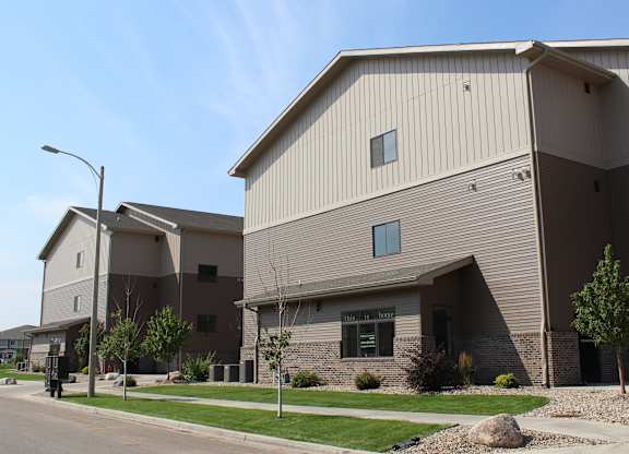 A contemporary apartment building exterior here at Dakota Commons showcasing a multi-story structure with neutral two-tone siding, brick accents along the base, landscaped greenery, sidewalks, and a quiet residential street under a clear blue sky.