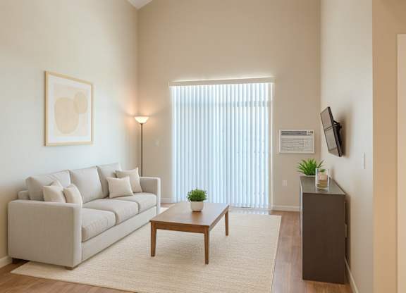 A bright, modern living room here at Dakota Ridge featuring high ceilings, a light neutral sofa with coordinating pillows, a wooden coffee table on a soft area rug, warm wood-style flooring, vertical blinds allowing natural light, a wall-mounted TV, and minimal decor creating an open, inviting atmosphere.