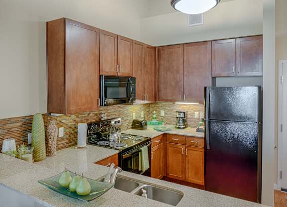 A warm, modern kitchen here at Dakota Ridge featuring rich wood cabinetry, light speckled countertops, a mosaic tile backsplash, black appliances including a refrigerator and microwave, under-cabinet lighting, and a functional peninsula with a stainless steel sink and simple decorative accents.