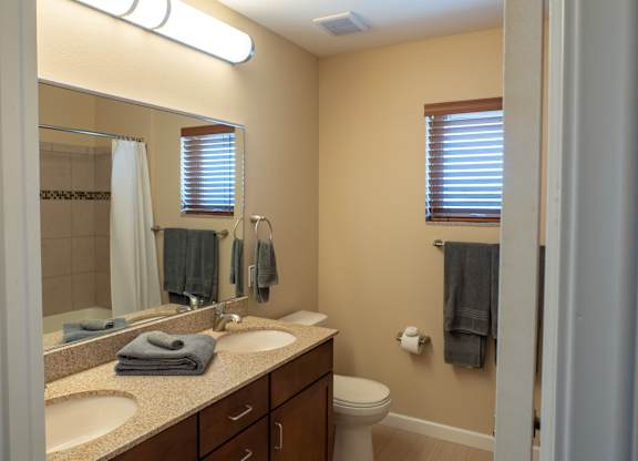A clean, well-lit bathroom here at Dakota Ridge featuring a dual-sink vanity with granite countertops, dark wood cabinetry, a large wall mirror, neatly folded towels, a toilet, neutral wall tones, wood-style flooring, and a small window with blinds that brings in natural light.