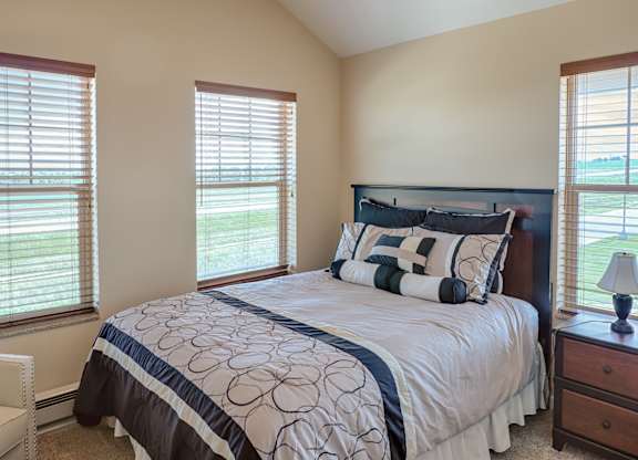 A bright, inviting bedroom here at Dakota Ridge featuring a neatly made bed with patterned bedding, a dark wood headboard, matching nightstands with a table lamp, soft carpet flooring, tall windows with wood blinds that bring in natural light, and neutral walls that create a calm, comfortable atmosphere.