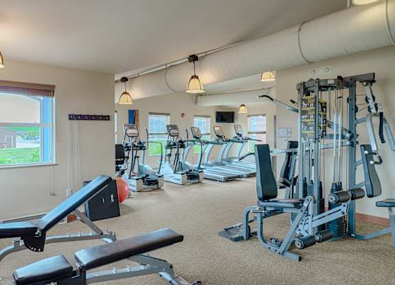 A spacious, well-lit fitness center here at Dakota Ridge featuring treadmills lined along large windows, a multifunction weight machine, a padded workout bench, neutral carpet flooring, exposed ceiling ductwork, and warm pendant lighting that creates a clean, motivating environment for residents to exercise comfortably.