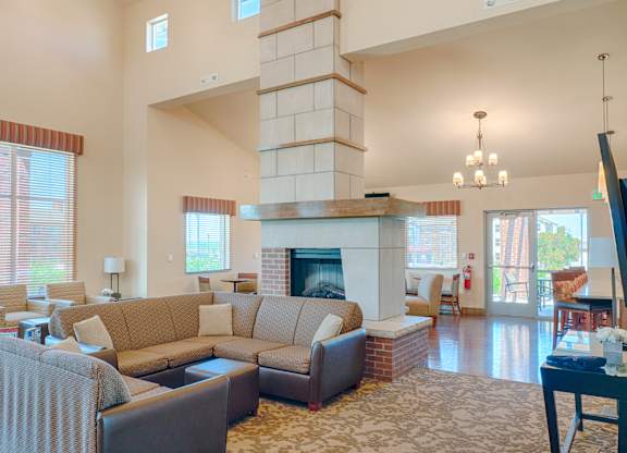 A welcoming community lounge here at Dakota Ridge featuring a large stone fireplace with brick accents, cozy sectional seating with neutral cushions, wood flooring, tall windows that bring in natural light, soft area rugs, and nearby tables and chairs that create a comfortable space for relaxing or gathering with neighbors.