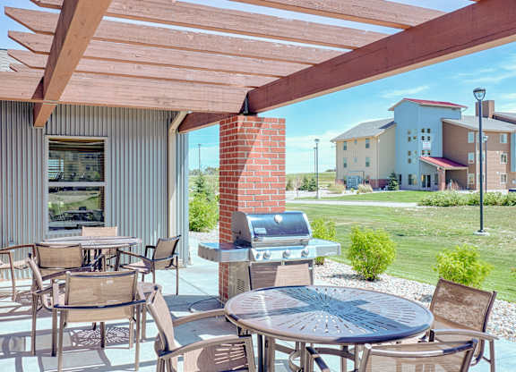 A shaded outdoor patio here at Dakota Ridge featuring a wooden pergola with exposed beams, round metal tables with mesh chairs, a stainless steel grill beside a brick column, and open views of manicured green lawns and nearby apartment buildings under a bright blue sky.