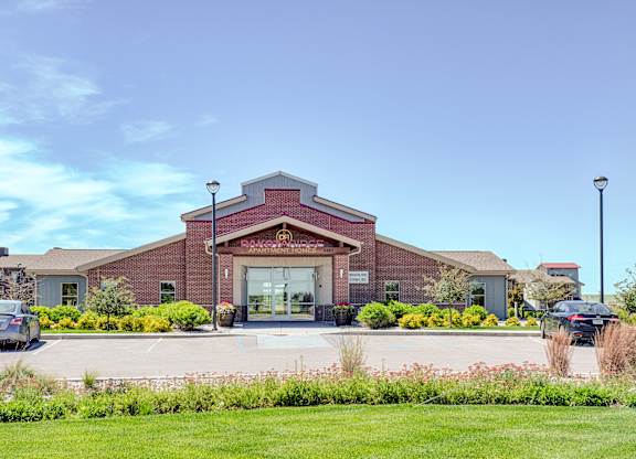A welcoming exterior view here at Dakota Ridge featuring the brick clubhouse entrance with a peaked roofline, glass double doors, landscaped greenery, colorful planters, and a wide parking area set against a bright blue sky and well-maintained lawns.