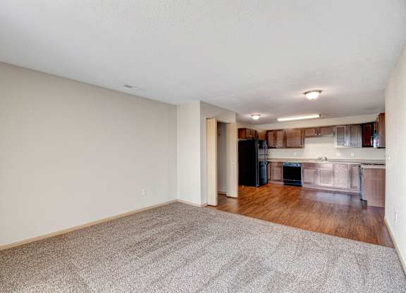 A large empty room with a carpeted floor and a kitchen area in the background.