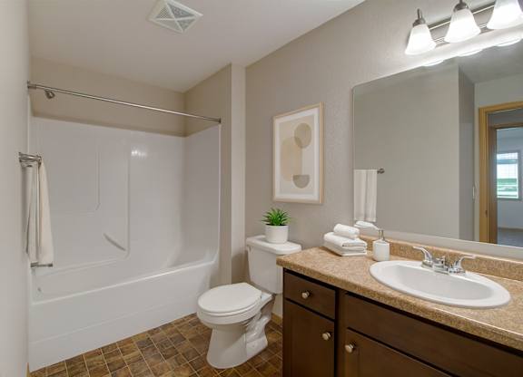 A clean bathroom here at Pheasant Ridge featuring a white tub and shower combination with smooth surround walls, a single-sink vanity with dark wood cabinetry and a neutral countertop, a large wall-mounted mirror with bright overhead lighting, folded white towels, simple wall art, and durable flooring in warm earth tones.