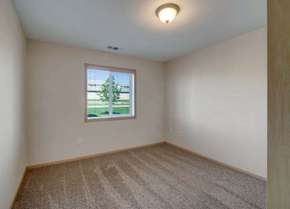 A bright bedroom here at Pheasant Ridge featuring plush neutral carpeting, soft beige walls with light wood trim, a centered window that brings in natural daylight and outdoor views, clean lines with ample wall space for furniture placement, and a simple ceiling light that creates a calm, comfortable atmosphere.