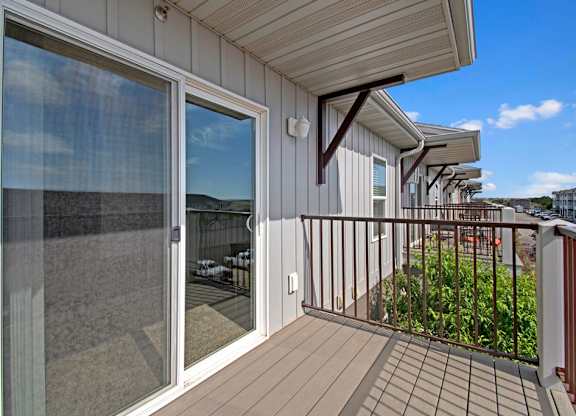 A private outdoor balcony here at Pheasant Ridge featuring durable wood-style decking, a sliding glass door with large glass panels, neutral gray siding, and a sturdy metal railing overlooking landscaped grounds and neighboring buildings under a bright blue sky.