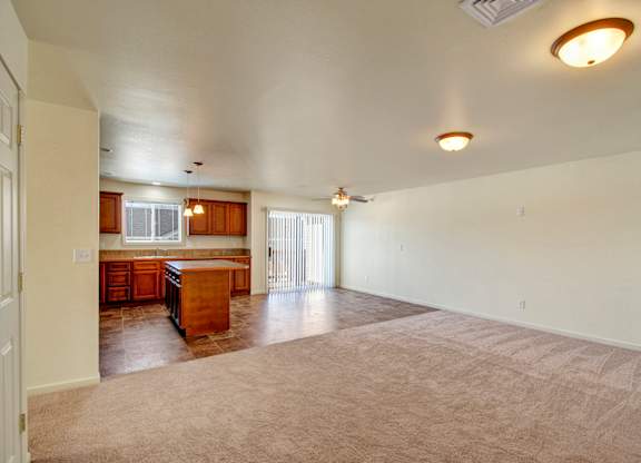 A spacious open living area here at Phoenix Ridge featuring soft neutral carpeting, an open-concept layout flowing into a kitchen with warm wood cabinetry, a center island, tile flooring, pendant lighting, and a sliding glass door that brings in natural light and connects to an outdoor space.