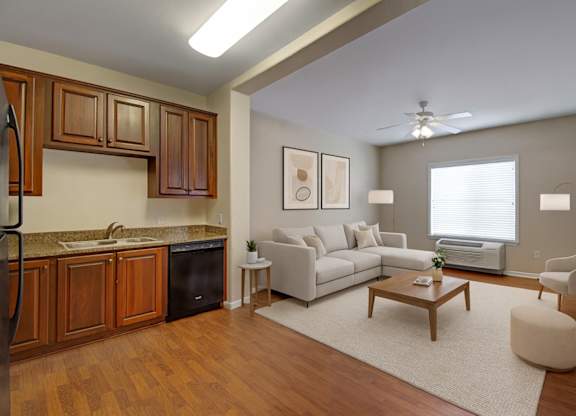A bright open living room here at Prairie Vista featuring warm wood-look flooring, a modern neutral sectional sofa, coordinating accent chairs, a wooden coffee table, soft area rug, and an adjacent kitchen with rich cabinetry, creating an inviting and functional apartment layout.