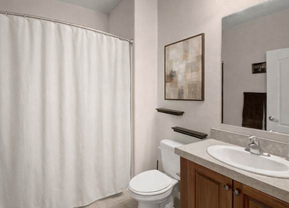 A clean bathroom here at Prairie Vista featuring a single-sink vanity with a light countertop, wood cabinetry, a large wall mirror, a toilet, floating wall shelves, neutral wall art, and a shower-tub combo with a white curtain and light tile flooring.