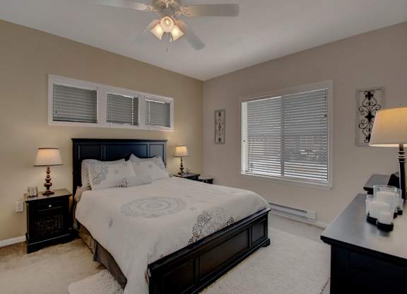 A cozy bedroom here at Prairie Vista featuring a dark wood bed with light patterned bedding, matching nightstands with table lamps, a ceiling fan with light, soft carpet flooring, neutral walls, decorative wall accents, a dresser, and windows with white blinds allowing natural light into the space.