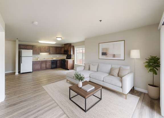 A bright, modern living room here at Renaissance Heights featuring light wood-style flooring, a neutral sectional sofa with plush pillows, a simple coffee table on a soft area rug, abstract wall art, a floor lamp, indoor greenery, and an open kitchen with dark cabinets and white appliances in the background.