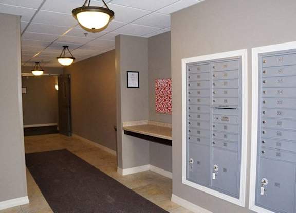 A clean, well-lit interior mailroom here at Renaissance Heights featuring organized wall-mounted mailboxes, neutral-toned walls, tiled flooring, overhead pendant lighting, and a convenient countertop area within a quiet residential hallway designed for easy mail access.