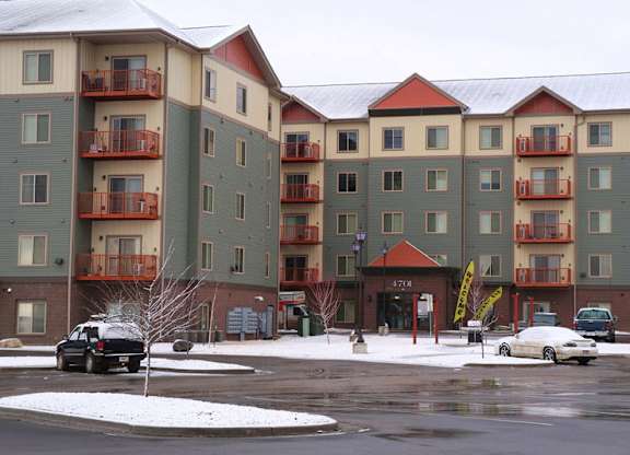 A snowy exterior view here at Renaissance Heights showcasing a multi-story apartment building with neutral siding, red-accented balconies, a covered main entrance, parked vehicles, and a paved parking area set against a winter landscape under an overcast sky.