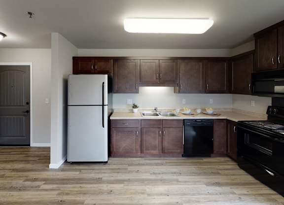 A spacious, modern kitchen here at Renaissance Heights featuring warm wood-style flooring, dark wood cabinetry, light countertops, a stainless steel refrigerator, black appliances, a double sink, and ample counter space with soft overhead lighting that creates a clean and functional cooking area.