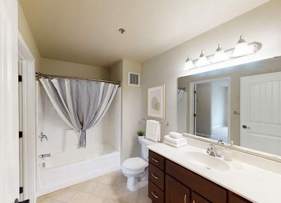 A clean, modern bathroom here at Renaissance Heights featuring a white tub and shower with a neutral curtain, a wide vanity with dark wood cabinetry and a white countertop, a large wall mirror with bright overhead lighting, light tile flooring, and neatly arranged towels that create a fresh and comfortable bathroom space.