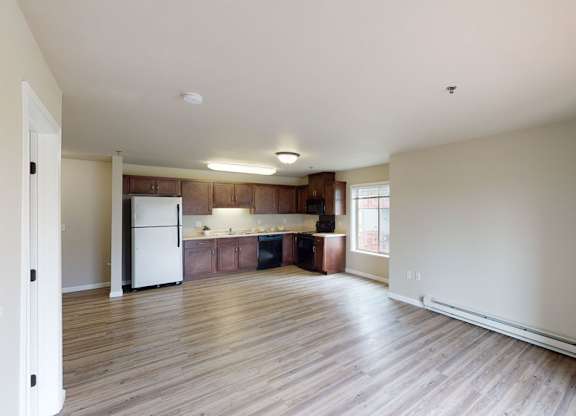 A spacious open-concept living room here at Renaissance Heights featuring wide plank wood-style flooring, neutral painted walls, and a bright layout that flows into a modern kitchen with dark wood cabinetry, light countertops, full-size appliances, and a large window that brings in natural light.