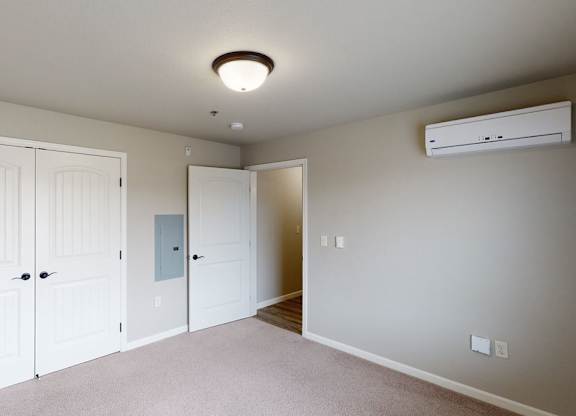 A bright and comfortable bedroom here at Renaissance Heights featuring soft neutral walls, plush carpet flooring, double white closet doors, a ceiling-mounted light fixture, and a wall-mounted mini-split unit, with an open doorway leading toward the living area for a clean and functional layout.