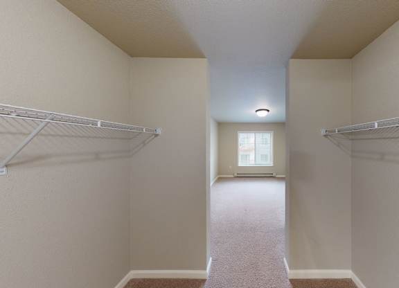 A spacious walk-in closet here at Renaissance Heights featuring dual white wire shelving on both sides, neutral walls, plush carpet flooring, and a hallway-style layout that opens toward a bright bedroom with a window and ceiling light.