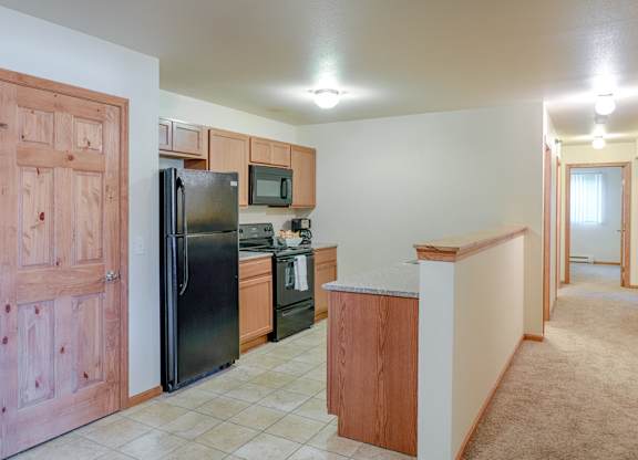 A warm open kitchen here at Rockridge featuring natural wood cabinets, black appliances including a refrigerator, range, and microwave, light tile flooring, a breakfast bar with wood trim, and an adjacent carpeted hallway that adds function and flow to the apartment layout.