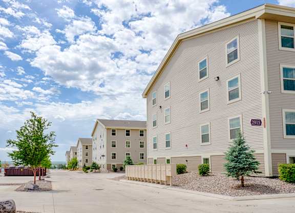 A well-maintained apartment community exterior here at Rockridge Apartments featuring beige multi-story buildings with white trim and multiple windows. A paved drive runs through the property, lined with mailboxes, landscaped rock beds, and small trees, creating an open, orderly setting under a bright, partly cloudy sky.