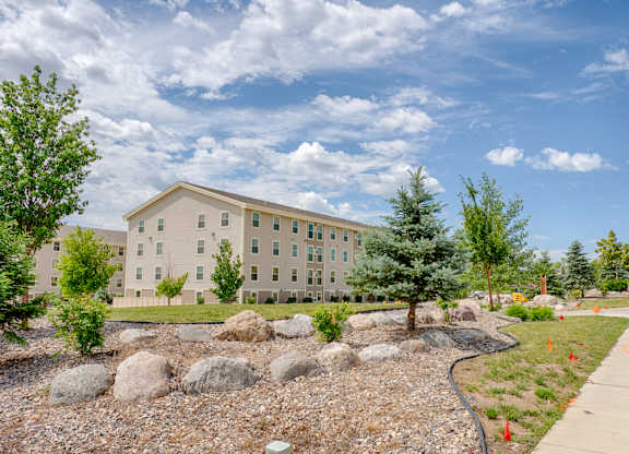 A landscaped outdoor view here at Rockridge Apartments featuring a multi-story beige apartment building set behind manicured green lawns, decorative rock beds, and mature trees. Large stones and greenery line the walkway, creating a peaceful, well-kept community setting beneath a bright, partly cloudy sky.