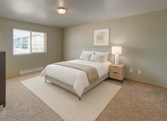 A bright, comfortable bedroom here at Vue 28 Apartments with beige carpet, soft neutral walls, and a large window with blinds that brings in natural light. The room features a neatly made bed with light bedding, a bedside table with a lamp, simple wall art, and an open layout that feels calm and inviting.