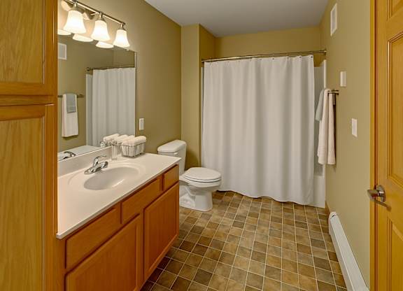 A clean, well-lit bathroom here at Vue 28 Apartments with tiled flooring, neutral walls, and a bathtub with a shower curtain. A wooden vanity with a white countertop and sink sits beneath a large wall mirror with overhead lighting, while a white toilet and towel bars complete the simple, functional layout.