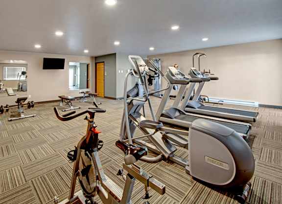 A well-equipped fitness center here at Vue 28 Apartments featuring cardio machines, weight equipment, and exercise benches arranged across a spacious, open floor plan. The room includes mirrored walls, wall-mounted televisions, patterned flooring, and bright overhead lighting, creating a comfortable and functional space for daily workouts.