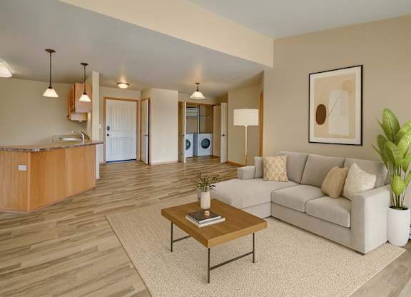 A bright, open living room here at Williston Garden Apartments with wood-style flooring, soft neutral walls, and an inviting layout that flows into the kitchen area. A light-colored sectional sofa sits on an area rug with a wooden coffee table, complemented by pendant lighting, simple wall art, and an in-home laundry area visible in the background.