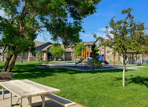 A lush, park-like outdoor green space here at Williston Garden Apartments featuring manicured lawns, mature trees, and a colorful playground set. A nearby picnic table and walking paths create a welcoming area for outdoor relaxation and recreation, with surrounding apartment buildings completing the peaceful community setting.