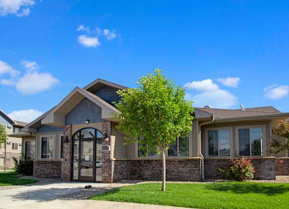 A welcoming community clubhouse here at Williston Garden Apartments with a brick and siding exterior, large windows, and a central glass entrance. Manicured lawns, young trees, and a paved walkway surround the building, creating a polished, inviting setting under a bright blue sky.