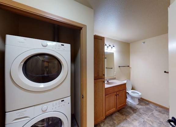 A convenient in-home laundry and bathroom area here at Williston Garden Apartments featuring a stacked washer and dryer tucked into a closet beside a bathroom vanity. The bathroom includes a wooden cabinet with sink, a white toilet, tiled flooring, and overhead lighting, offering a functional layout with everyday essentials close at hand.