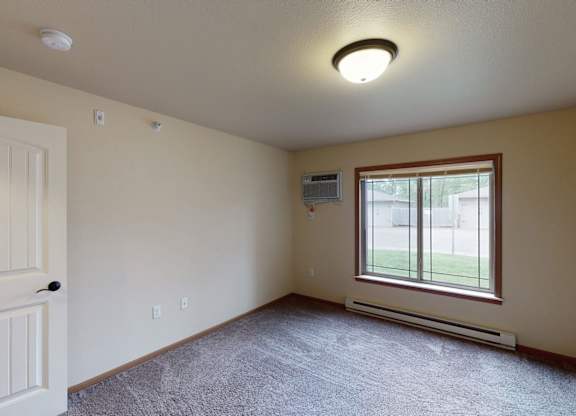 A bright, empty bedroom here at Williston Garden Apartments with plush carpet, soft neutral walls, and a large window that brings in natural light and outdoor views. A wall-mounted air conditioning unit, baseboard heating, and an open layout provide comfort and flexible space for bedroom furniture.