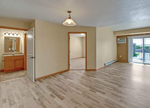 A spacious, open living area here at Williston Garden Apartments with wood-style flooring, soft neutral walls, and an airy layout. A sliding glass door with vertical blinds leads to a private balcony and allows natural light to fill the room, while baseboard heating, overhead lighting, and nearby access to the bedroom and bathroom add comfort and convenience.