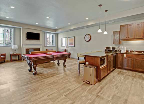 A stylish community lounge here at Williston Garden Apartments with wood-style flooring, recessed lighting, and a welcoming open layout. A pool table anchors the seating area near a wall-mounted television and fireplace, while a full kitchen with wood cabinets, an island, and pendant lighting creates a comfortable space for gatherings and social events.
