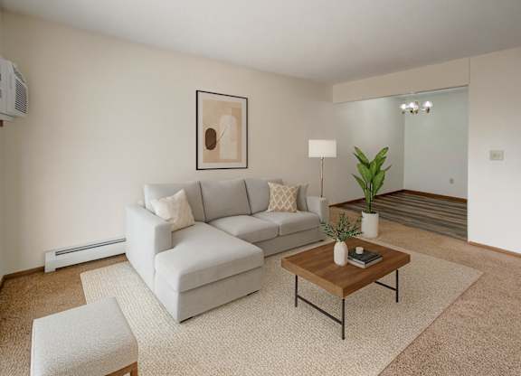 A cozy living room here at Windscape Apartments featuring plush carpet, soft neutral walls, and a modern sectional sofa arranged on a textured area rug. A wooden coffee table, simple wall art, and potted greenery add warmth, while an open view into the dining area creates a comfortable, inviting layout.