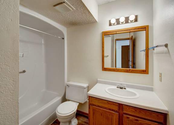A clean, functional bathroom here at Windscape Apartments with a tub and shower combination, neutral walls, and wood-finish vanity cabinetry. A wide mirror with overhead lighting, durable flooring, and a simple layout create a practical, comfortable space designed for everyday use.